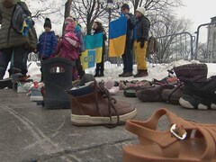 Des souliers d'enfants posés sur le sol devant l'Assemblée nationale. Il y a de la neige par terre et dans le fond des gens qui tiennent le drapeau ukrainien dans leurs mains.