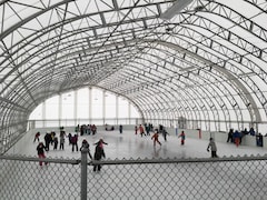 Des enfants patinent sur une patinoire protégée. La structure est faite d'acier ou aluminium  et le recouvrement en toile blanche qui laisse passer une partie de la lumière.