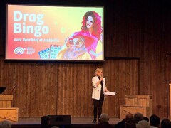 Monique Poirier sur scène devant un public au Pays de la Sagouine. Derrière elle, un immense écran qui porte la mention drag bingo avec Rose Beef et Joséphine.