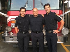 Un père et ses deux fils se tiennent bras dessus, bras dessous en uniforme de pompier devant leur camion. 