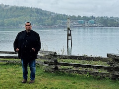 Rueben George pose sur un rivage. Au loin on voit les quais et les réservoirs d'un terminal pétrolier.