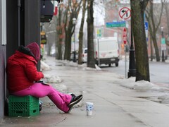 Une femme habillée en rose, un capuchon sur la tête, est assise sur une caisse de lait avec un gobelet devant elle, sur le trottoir d'une rue déserte à la fin de l'hiver.