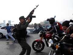 Un soldat pointe son arme dans les airs dans une rue où des civils courent et des hommes se déplacent à moto.