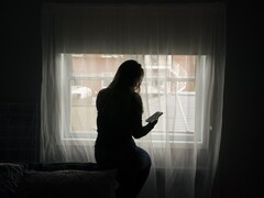 Une femme consulte son téléphone cellulaire assise dans une pièce plongée dans la pénombre, devant une fenêtre d'un quartier résidentiel.