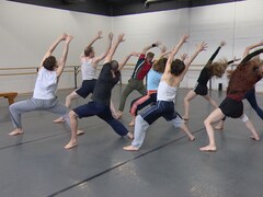 11 danseurs exécutant un mouvement les bras dans les air dans une salle de ballet au plancher gris et murs de briques blancs. Mai 2024.