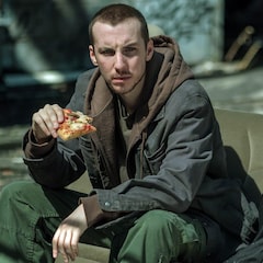 Jessy est assis sur un vieux siège de fourgonnette, dans un lieu abandonné et il mange une pizza.