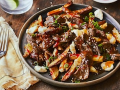 Une poutine recouverte de bulgogi de bœuf est dans une assiette, accompagnée d’une sauce au gochujang, d’oignons verts et de graines de sésame.