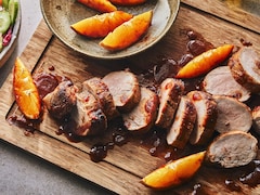 Sur une planche de bois, un filet de porc est coupé en tranches avec des quartiers d’orange et un filet de sauce au piment chipotle.