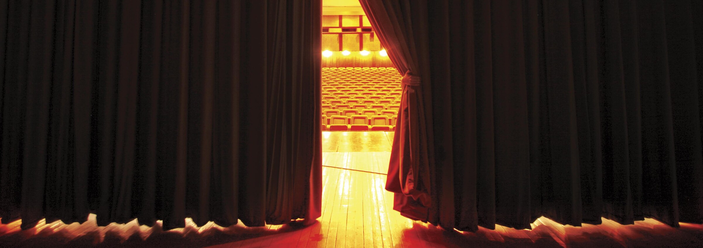 Un rideau de théâtre rouge, entrouvert montre la salle éclairée et des sièges vides.