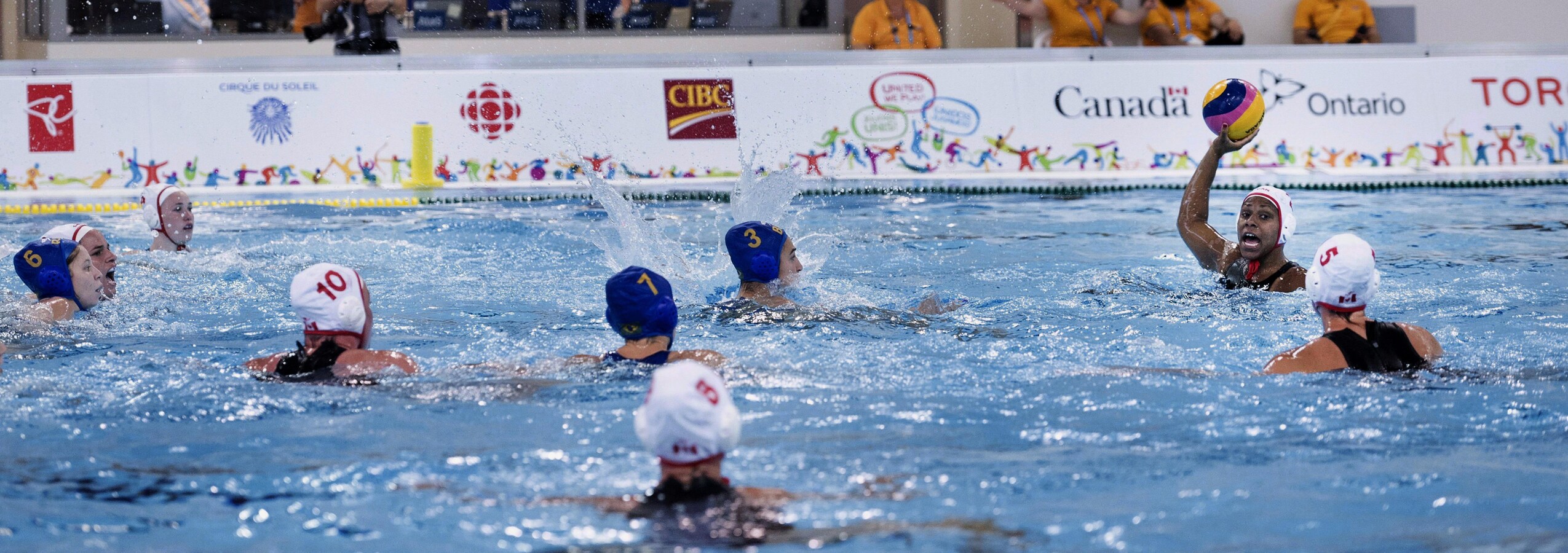 Une joueuse de water-polo tient le ballon haut dans les airs et regarde d'autres joueuses qui nagent sur place.