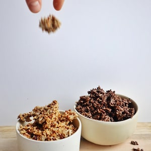 Sur une table, deux bols remplis de granolas préparés de deux façons, et une main laissant tomber un morceau de granola vers un des bols.