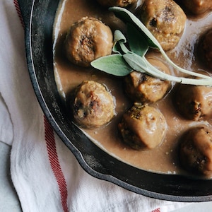 Vue en plongée sur une demi-poêle en fonte contenant des boulettes baignant dans une sauce et garnies de feuilles de sauge.