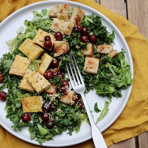 Dans une assiette, il y a une salade verte garnie de morceaux de tofu grillés, des raisins et des croûtons.