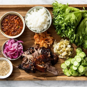 Un plateau en bois dans lequel se trouve du porc à la coréenne, de la laitue frisée, des tranches de concombres libanais, du riz, des graines de sésame et du kimchi.