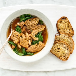 Un bol contenant une soupe aux saucisses et haricots blancs. Des croutons au parmesan sont disposés à la droite du bol de soupe.
