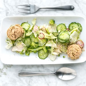 Une salade de légumes dans une assiette de service avec deux boules de thon avec à ses côtés un bol de vinaigrette verte.