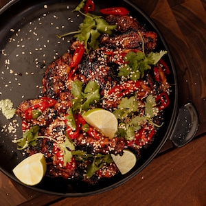 Dans un plat en fonte, il y a des ailes de poulet grillées garnies de graines de sésame, de coriandre fraîche et de quartiers de lime.
