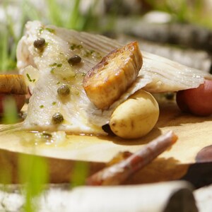 Un morceau d'aile de raie cuite, couverte de beurre aux câpres, accompagnée de pommes de terre, et déposée sur une planche en bois.