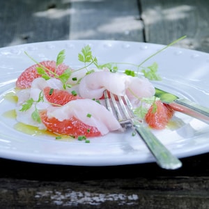 De fines tranches de poisson blanc et des suprêmes de pamplemousse rose parsemés d'herbes fraîches, dans une assiette blanche.