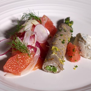 Un morceau de saumon recouvert de morceaux de pamplemousse, de radis et d'aneth avec une petite crêpe roulée avec des asperges à l'intérieur et de la crème fraîche au raifort.
