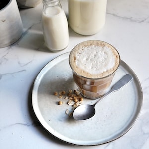 Un verre de boisson soya et avoine bien moussée pour le café, dans une assiette, à côté de grains de soya et d'avoine, et d'une cuillère à café.