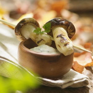 De la crème sure dans un petit bol en terre cuite et une brochette en bois de deux champignons entiers et rôtis disposée sur le dessus.
