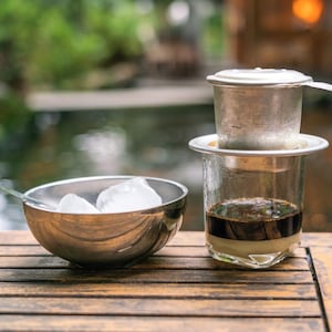 Un verre contenant du lait condensé et du café infusé, un filtre à café vietnamien posé sur le dessus du verre et un bol de glace.