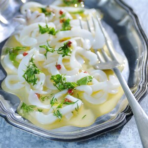 Dans un plat en argenterie, des calmars en fines tranches de calmars baignent dans une sauce crémeuse jaune parsemée de persil.