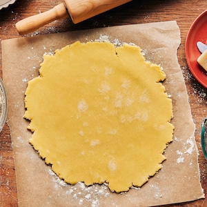 Une pâte à tarte sans gluten est abaissée sur un papier parchemin et accompagnée d’un bol de farine, d’une tasse à mesurer, de beurre et d’un rouleau à pâtisserie.