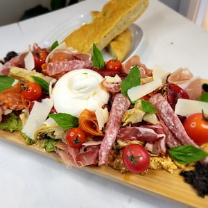 Une planche de présentation garnie de tranches de charcuteries, de légumes marinés, une boule de burrata et des copeaux de parmesan.