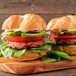 Deux hamburgers au porc sur une planche à découper. Ceux-ci sont garnis de tranches d'avocat, de tranches de tomate, des épinards, ainsi que des oignons rouges.