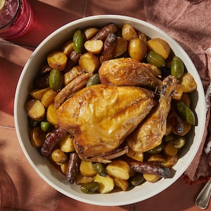 Dans une assiette de présentation, il y a un poulet rôti avec des saucisses merguez, des pommes de terre rôties et des olives vertes.