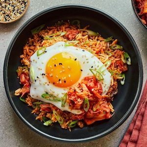 Dans un bol noir, un œuf est déposé sur du riz frit au kimchi. Autour, on voit des graines de sésames, des oignons verts et un petit bol de kimchi.