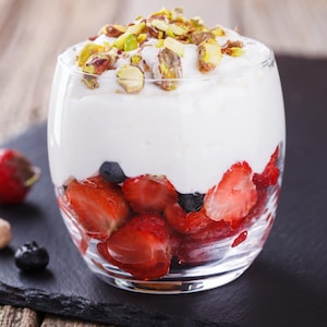 Un verre contenant des fraises. Elles sont surmontées de crème chantilly au chocolat blanc et de pistaches rôties.