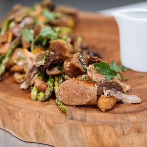 Sur une planche à découper, il y a des saucisses de chevreuil à l'érable avec des asperges et des champignons en accompagnement.