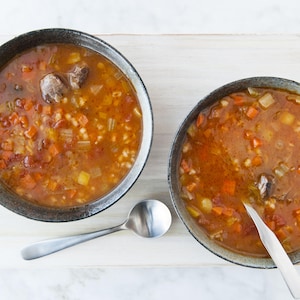 Des bols de soupe au boeuf et à l'orge servis sur une planche de bois, avec des cuillères à soupe.