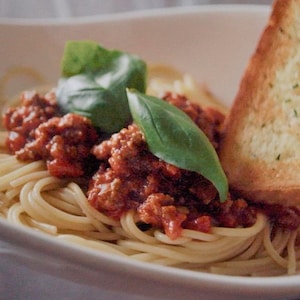 Un bol de spaghetti, garni de sauce bolognaise et de basilic frais, accompagné d'une tranche de pain à l'ail.