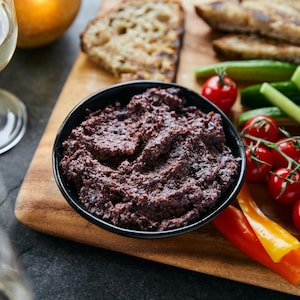 Bol de tapenade aux olives Kalamata sur une planche de bois avec des légumes, du pain grillé et une coupe de vin blanc.