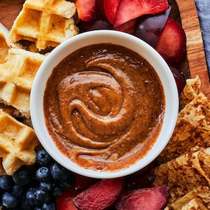 Au centre de la photo, est déposé un bol contenant la trempette au café et au beurre d'amandes. À l'entour, il y a des bleuets, crêpes, gaufres et prunes. À la droite du bol, il y a deux fourchettes sur une serviette de table. 