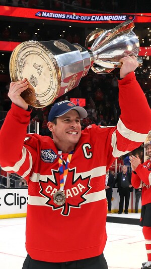 Sur une patinoire, un joueur de hockey tient un trophée à bout de bras devant d'autres joueurs souriants et des partisans contents.