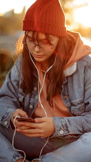 Une adolescente portant un jean, un manteau de jean, une tuque et des lunettes est assise par terre dans la nature en automne. Elle texte sur son téléphone et écoute de la musique dans des écouteurs avec un fil blanc.