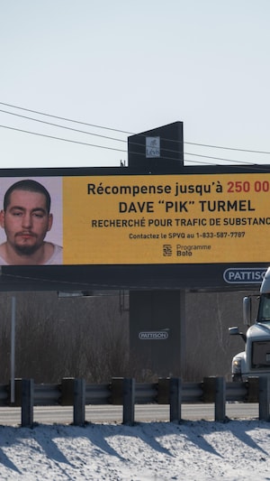 Un panneau d'affichage avec la photo du fugitif Dave Turmel. Un camion semi-remorque circule sur l'autoroute.
