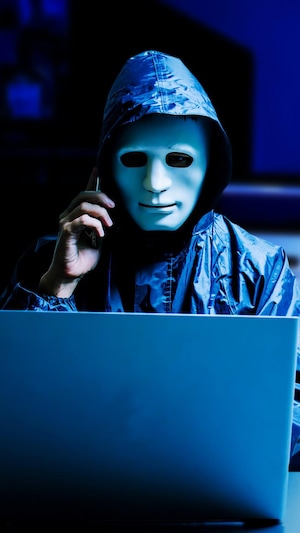 Un pirate informatique portant un masque parle au téléphone devant un ordinateur portable.