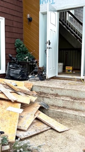 Un tas de planches de bois moisies et des objets abîmés, devant les marches d'une maison, dont la porte principale est ouverte