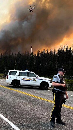 Un policier et sa voiture sur une route, un imposant nuage de fum&eacute;e s'&eacute;l&egrave;ve de la for&ecirc;t derri&egrave;re lui avec un h&eacute;licopt&egrave;re.