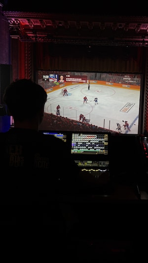 Un homme derrière une console regarde un match de hockey diffusé dans une salle de spectacle. 