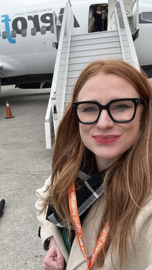 Julie-Anne Lapointe prend un égoportrait sur le tarmac d'un aéroport, on voit un avion derrière elle.