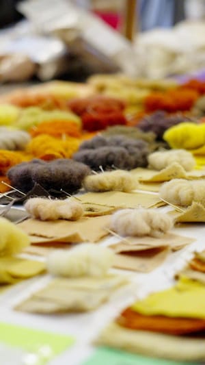 Un plateau sur lequel reposent des carrés de tissus et des pelottes de laines colorés en tons orangés, jaunes, bruns et noirs.