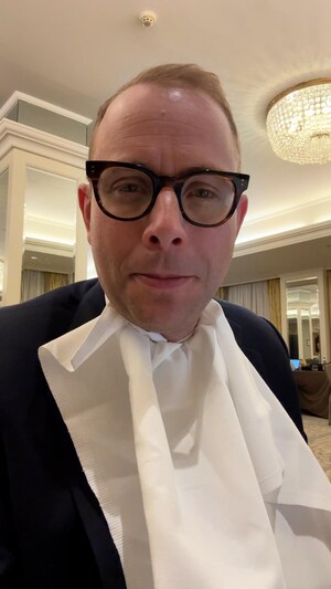 Louis Blouin est assis dans une salle de conférence, devant son ordi. Il porte un bavette afin d'éviter de salir sa chemise en mangeant tout en terminant un reportage.