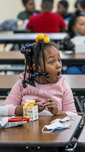 Deux petites filles sont assises dans la cafétéria d'une école pour manger. L'une montre quelque chose qu'elle a dans son lunch et l'autre a la bouche grande ouverte, car elle est émerveillée de ce qu'elle voit.